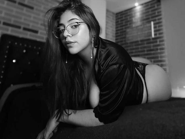 RousseColliinss with glasses, long hair, and a sexy look, wearing a leather shirt and lace panties, on all fours, showing her big ass and cleavage in black-and-white.