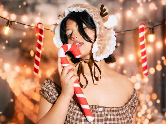 MisakiWong with black hair, eyes closed, wearing a furry reindeer hat and brown checkered dress, licking a red and white candy cane, against a warm, blurred light background.