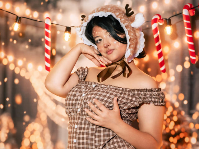 MisakiWong with dark hair, wearing a cute reindeer hat, brown plaid off-shoulder top, touching her chin and tits, surrounded by warm fairy lights and candy cane decorations.