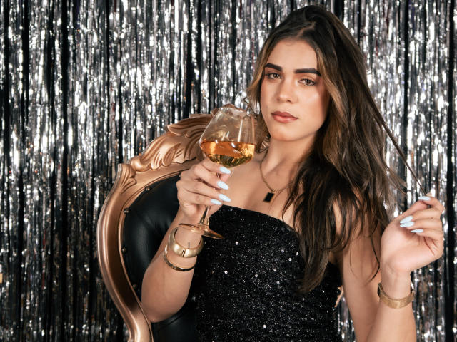 AliceCoolenk with long, wavy brown hair sits in a gold, ornate chair holding a glass of amber liquid. She wears a black, glittery strapless dress, gold bracelets, and a black necklace. Her light blue nails stand out against the gold and black. She has a confident, slightly relaxed expression. The background is a silver, reflective curtain. The composition focuses on her and the glass, highlighting the contrast between her dark dress and the shiny curtain.