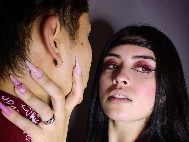 NoahAndRose, com maquiagem rosa, tranças pretas e unhas longas, tocando suavemente o rosto de outra pessoa. Nariz visível, lábios rosados e um anel na mão de NoahAndRose. Imagem íntima, em close-up..