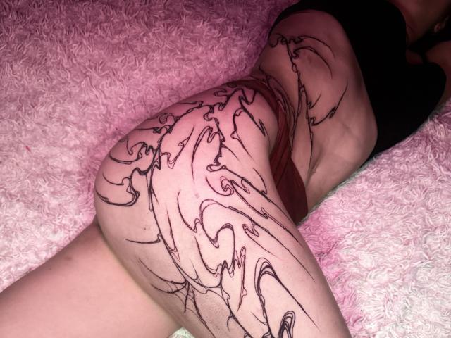 EnaMore's tattooed ass, lying on a pink, fluffy surface, with black, swirling lines covering her lower back and right thigh, wearing a black top, in a slightly angled shot.
