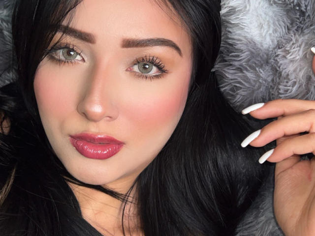 LillyTaylor's face with glossy pink lips, green eyes, black hair, white nails, and a slight pout, lying on a fluffy gray surface.