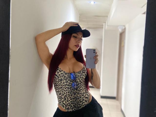 ValeryHarllow with red hair, wearing a black cap, leopard print top, and black pants, showing her large tits and holding a phone in a hallway.