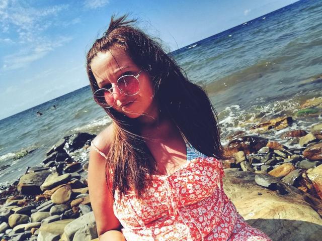 LeonoreElly with wet brown hair, wearing round sunglasses and a red floral dress, standing on rocky shore, blue ocean in background, sunlight, slight smile, small breasts.