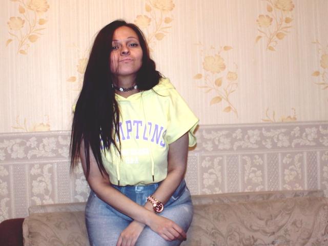LeonoreElly with long black hair, yellow shirt, blue jeans, sitting on a couch, smiling, wearing a pink watch, necklace, and a slight pout, looking cute and confident.