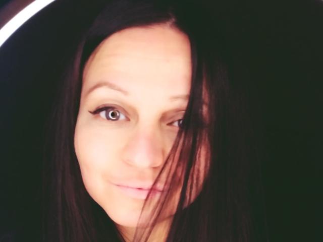 Close-up portrait of LeonoreElly with light skin and brown hair partially covering her face. She has brown eyes with subtle eyeliner, a slight smile, and a relaxed expression. Her hair is straight and falls over her right eye. The image has a soft, slightly blurred effect, with a circular white highlight in the upper left. The overall composition focuses on LeonoreElly's face, emphasizing her gentle expression and natural beauty.