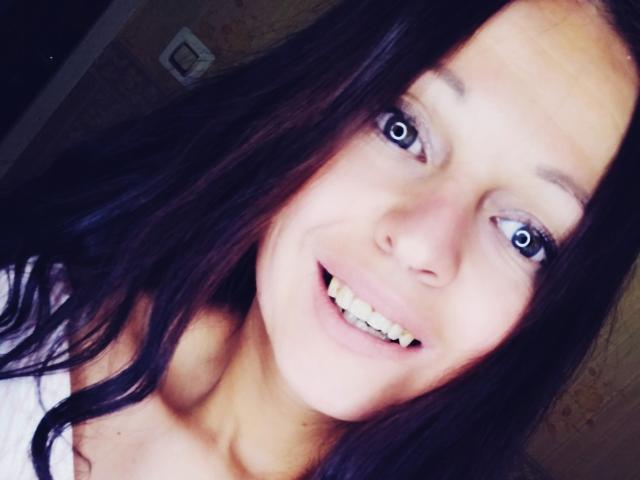 Close-up selfie of LeonoreElly with bright blue eyes, slightly parted lips showing teeth, and a subtle smile. LeonoreElly has long, dark brown hair, fair skin, and a slight blush on her cheeks. She's wearing a white top with a low neckline. The image is slightly blurred, emphasizing her eyes and smile. The lighting is soft, highlighting her facial features. The overall composition is casual and intimate.