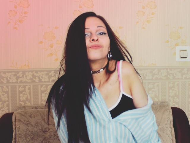 LeonoreElly with long black hair, blue eyes, wearing a striped shirt off-shoulder, black and pink bra, choker, and hoop earrings, sitting on a couch, smiling slightly.