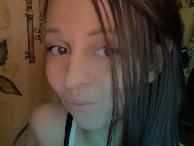 Close-up selfie of LeonoreElly with wet brown hair partially covering her face. She's winking with her right eye and puckering her lips. Her skin is light, and she has small, dark eyebrows. She's wearing a black and white strap top. The background is a yellow, patterned tapestry with an antique key design. The lighting is soft, and the image has a slightly blurry texture. LeonoreElly's expression is playful and casual.