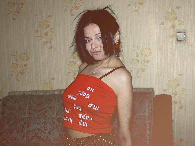 LeonoreElly has messy black hair, looking to the side with a slight smile. She wears a red strapless crop top with white text, exposing a brown bra strap. She's in a room with beige floral wallpaper and a patterned couch. She has a silver necklace and small earrings. Her skin is light and she has a slim build. The image has a slight color filter, giving it a warm, vintage look.