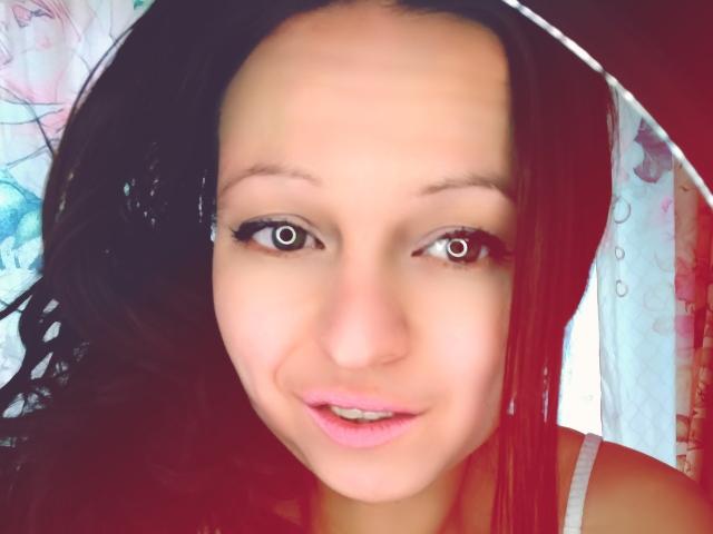 Close-up of LeonoreElly with light brown skin and dark brown hair. She has large brown eyes, subtle black eyeliner, and pink lipstick. Her hair is slightly wet and she wears a white headscarf with floral patterns. The background is blurred with pastel colors. LeonoreElly's expression is neutral with a slight smile. The image has a soft, slightly overexposed look.