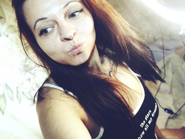 A close-up selfie of LeonoreElly with light skin and brown hair. She pouts her lips slightly to the side, wearing a black tank top with white text. Her eyes are looking to the left. Her hair is straight and falls over her right shoulder. She has a small, delicate nose and light makeup. The lighting is soft, creating a slight glow on her face. The image is slightly blurred.