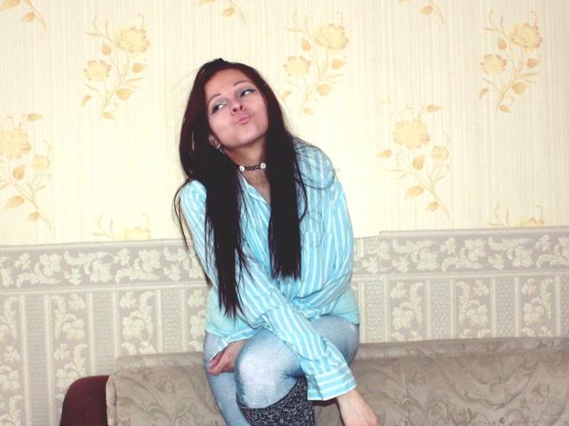 LeonoreElly, with long black hair, wearing a blue striped shirt and jeans, sits on a floral-patterned couch, looking slightly up with a playful expression, hands on knees.