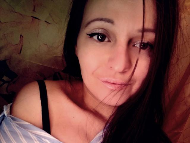 Close-up of LeonoreElly with medium-length dark brown hair, slightly wavy, partially covering her right eye. She has light skin, subtle makeup with black eyeliner, and a small, centered nose. She's wearing a light blue, off-shoulder shirt with a black strap visible on her left shoulder. Her expression is neutral, with a slight, almost invisible smile. The lighting is warm, highlighting her facial features. The texture of her hair is smooth, contrasting with the slightly rough texture of the background. Her eyes are brown, looking directly at the camera. The image has a casual, candid feel.