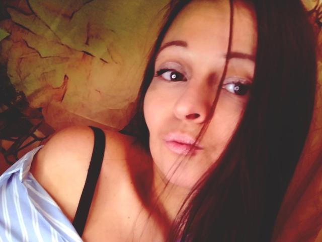 LeonoreElly with puffy pink lips, brown eyes, and long dark hair, wearing a blue shirt with a black strap, looking up with a slight pout. Boobs visible, soft light.