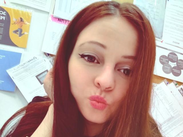 LeonoreElly with long brown hair, pouty pink lips, and winged eyeliner, making a kissy face, her hand in her hair, looking slightly to the side.