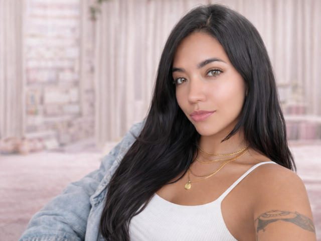 SofieWhite has long black hair, tan skin, brown eyes, pink lips, white tank top, gold necklace, tattoo on right arm, denim jacket, looking at camera, smiling subtly.