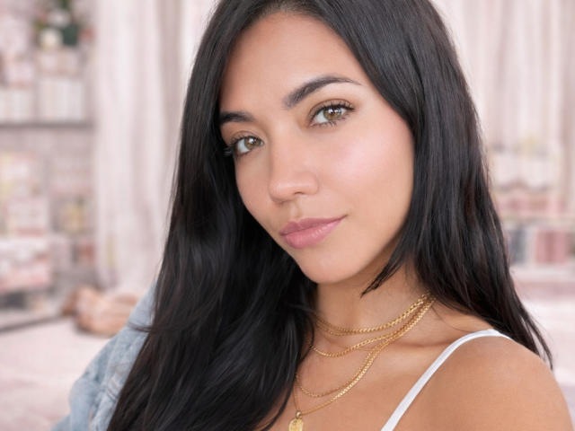 Close-up of SofieWhite with long, straight black hair and brown eyes. She has a light tan skin tone and wears pink lipstick. She's looking at the camera with a slight smile. She's wearing a white spaghetti strap top and multiple gold necklaces. The background is blurred, showing a cityscape. Her eyebrows are thick and well-groomed. Her makeup is subtle, highlighting her natural features. The image has a soft, realistic texture.