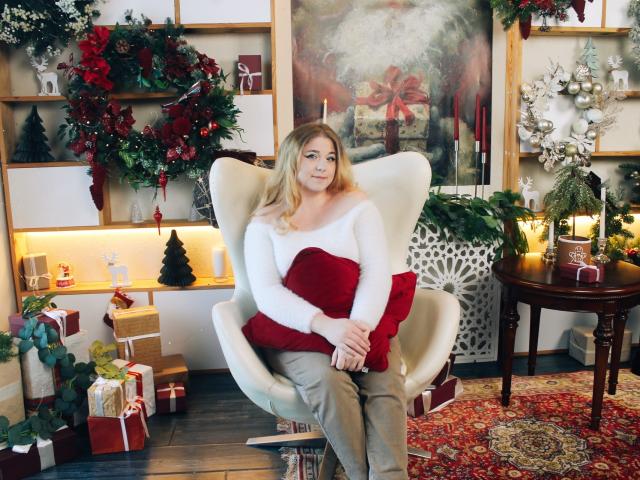 Jessika Daniels, con cabello rubio, blusa blanca de corte asimétrico y un cojín rojo en una silla color crema, tomándose de las manos, rodeada de adornos navideños, regalos y una mesa de madera..