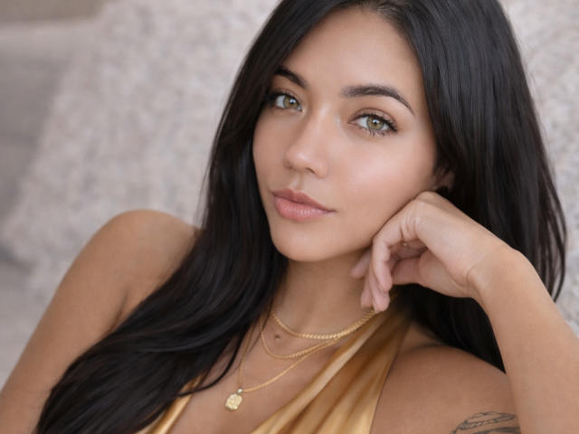 SofieWhite has long black hair, green eyes, and a small gold necklace with a heart pendant. She's looking at the camera with a slight smile, resting her chin on her hand.