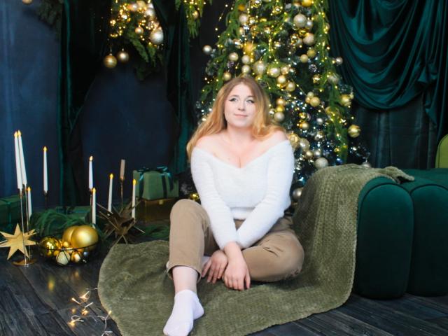 JessikaDaniels with blonde hair, white off-shoulder top, and beige pants, sitting on green blanket, wearing white socks, next to Christmas tree with gold ornaments and candles.