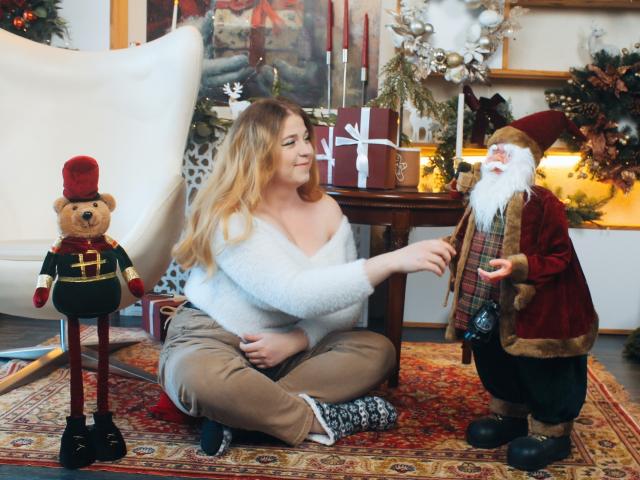 Jessika Daniels mit welligem, blondem Haar, einem weißen, flauschigen Oberteil und beigen Hosen sitzt auf einem roten Teppich und berührt eine Weihnachtsmann-Figur, während ein Teddybär in einem grünen Anzug in der Nähe steht..