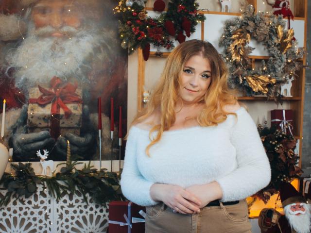 Jessika Daniels, con cabello rubio, un suéter blanco y suave, y pantalones beige de cintura alta, está de pie en una habitación decorada para Navidad, tomándose de las manos con alguien frente a ella, y sonriendo a la cámara..