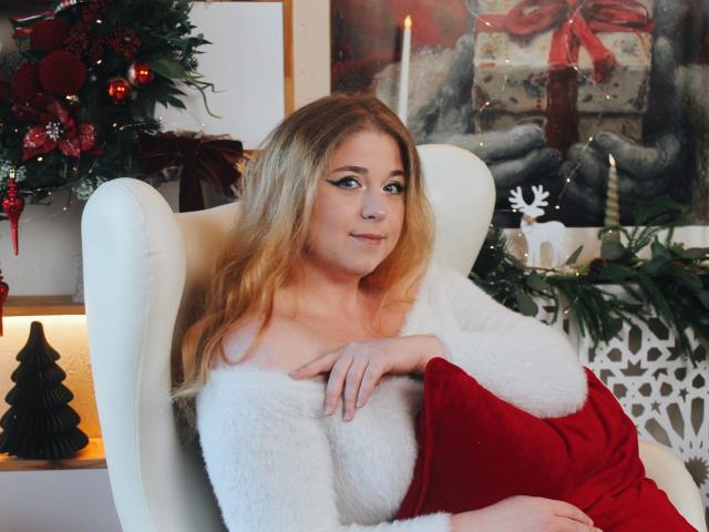 Jessika Daniels mit blonden Haaren, einem weißen Fellbademantel und einem roten Kissen, sitzt auf einem weißen Stuhl, sieht niedlich aus, hat ein sanftes Lächeln, ist mittelgroß gebaut, im Hintergrund sind Weihnachtsdekorationen, Kerzen und Geschenke zu sehen..