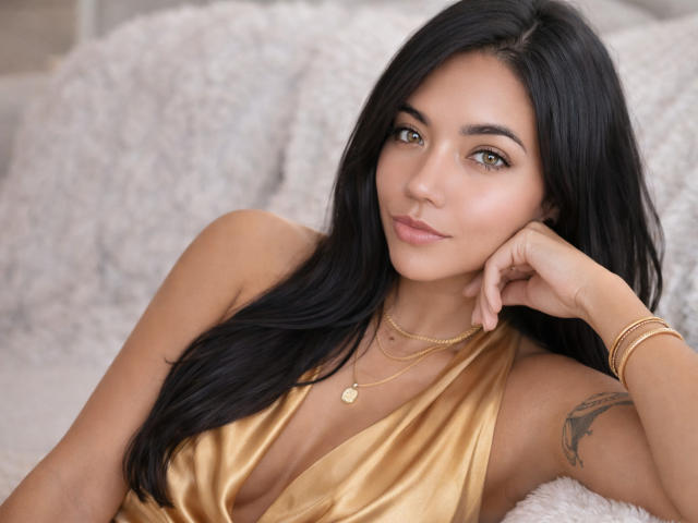 SofieWhite with long black hair, green eyes, and tan skin, wearing a gold dress, lying on a fluffy white surface, smiling, hand on cheek, gold necklace, and bracelet.