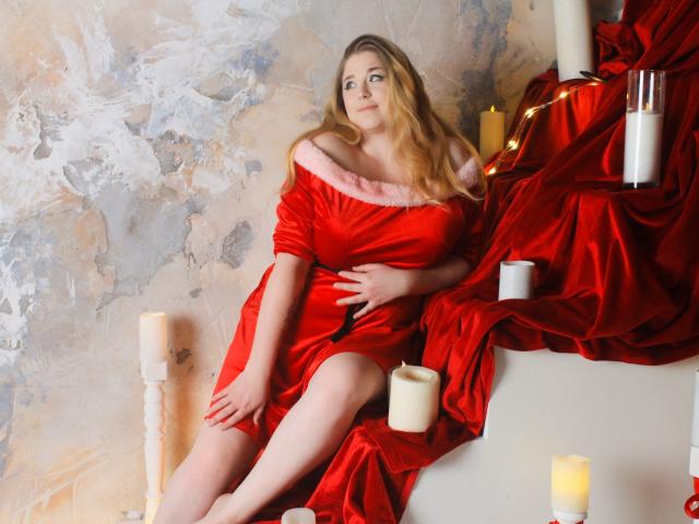 JessikaDaniels sits on a white staircase, wearing a red satin dress with pink fur trim. Her blonde hair is loose, and she looks to the side with a slightly pensive expression. Red satin drapes around her, and white candles are scattered on the stairs and in the background. The wall has a textured, unfinished look with gray and beige tones. The red satin contrasts sharply with the white candles and staircase. The image has a soft, warm light, enhancing the satin's shiny texture. This is a photograph with a focus on luxurious, romantic aesthetics.