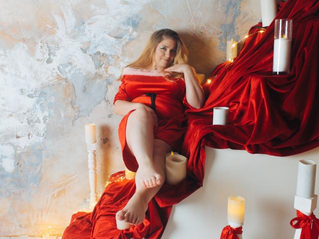 JessikaDaniels with red dress, sitting on white staircase, legs crossed, bare feet, candles around, red velvet draping, blonde hair, hand on chin, soft smile, relaxed, attractive.