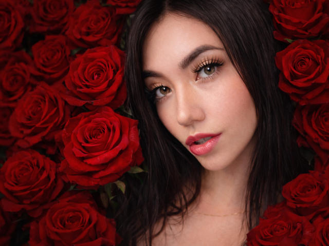 SofieWhite with dark hair, brown eyes, and pink lips, surrounded by red roses, looking directly at the camera, with a subtle gold necklace.