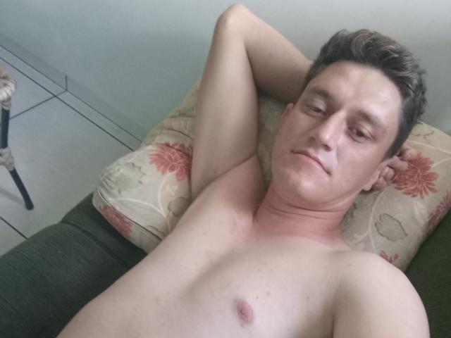 Eliss69, shirtless, lying on a floral pillow, arms behind head, slight smirk, short brown hair, light skin, small nipple, green couch, tiled floor, relaxed, casual.