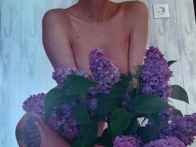 SophieCrach is topless with purple lilac flowers covering her tits and ass, holding a bouquet in front of her. Her arms are crossed, and a tattoo is visible.