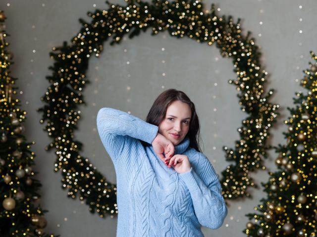 IsabelBlackberry with medium tits, wearing a light blue knitted sweater, smiling, hands near her chin, standing in front of Christmas wreaths with twinkling lights.