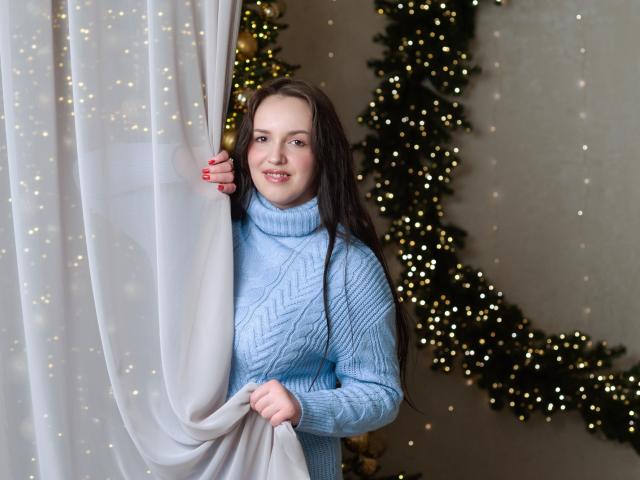 IsabelBlackberry with long brown hair, wearing a light blue turtleneck sweater, holding a white curtain, smiling, standing next to a Christmas tree with tiny glowing lights.