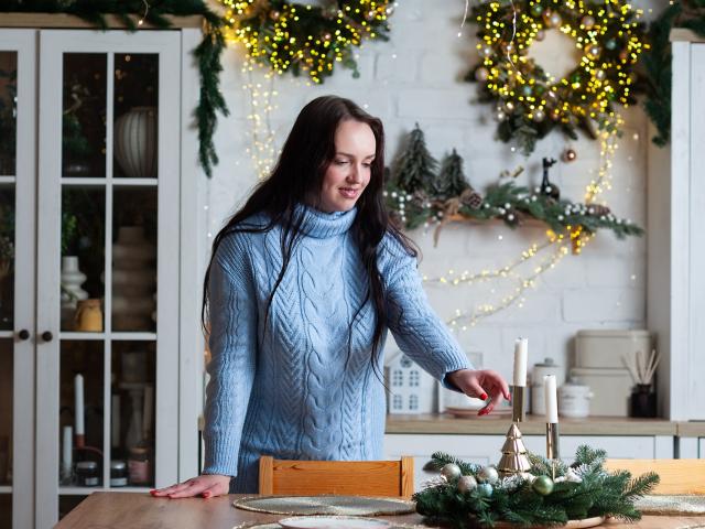 IsabelBlackberry stands in a cozy kitchen, wearing a light blue, chunky knit sweater with a cable pattern. She has long, straight brown hair and is smiling slightly as she reaches for a candle on a wooden table. The table holds a green wreath with pine branches and small ornaments. Warm, yellow fairy lights and Christmas decorations adorn the white brick wall behind her. IsabelBlackberry's right hand is on the table, and her left hand is reaching out. The overall composition is warm and festive.