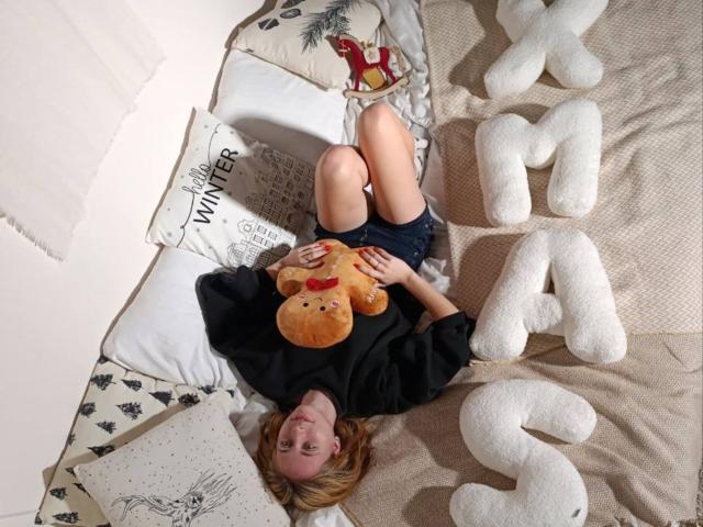 TracyLeex lying on a bed with white letters, wearing a black shirt and blue shorts, holding a teddy bear with a red nose, blonde hair, and a cute smile.