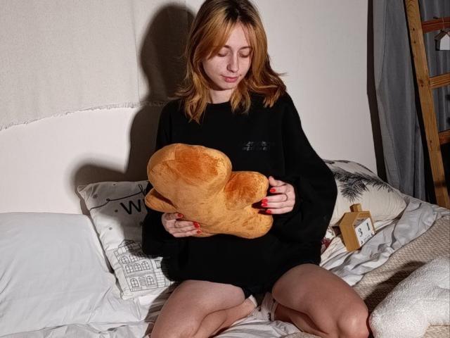 TracyLeex with medium brown hair, wearing a black shirt, holding a heart-shaped bread loaf, sitting on a bed, red nails, white pillows, and a wooden ladder in the background.