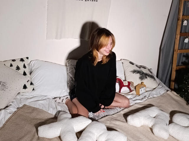 TracyLeex sits on a bed wearing a black oversized shirt, with blonde hair, small tits, and a cute smile, surrounded by white pillows and fluffy white socks.