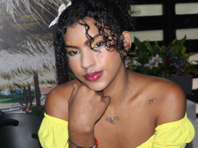SamaraDuvall with curly black hair, yellow off-shoulder top, pink lipstick, yellow eye makeup, small heart tattoo on right chest, hand on neck, dark skin, looking at camera.