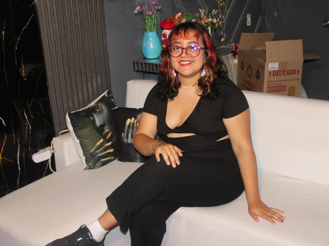 NamiNerona with red and black curly hair, glasses, black dress with cutouts, showing her tits, sitting on white couch, black and green pillow, cardboard box, and flowers in background.