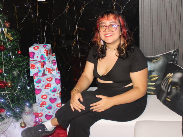NamiNerona, with red and black curly hair, wearing a black cut-out top and pants, sits on a white couch, smiling, with glasses, Christmas gifts, and a decorated tree.
