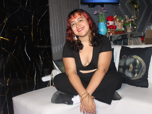 NamiNerona, a curvy Latina woman with red and black hair, sits on a white couch in a black and white outfit, smiling with her boobs slightly visible in a deep cut.