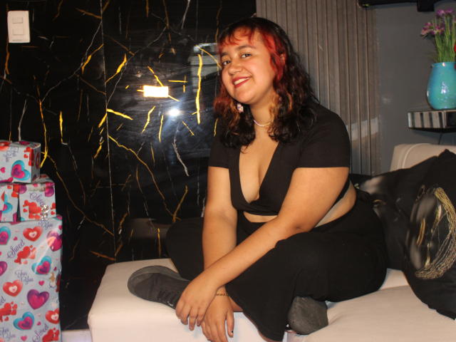 NamiNerona, curvy woman with red and black hair, sitting on white couch, black top revealing her boobs, black pants, smiling, gift boxes with hearts on left, black wall behind.