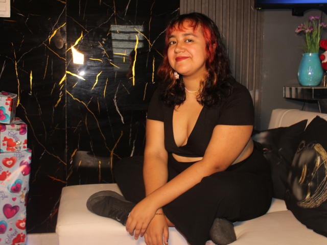 NamiNerona with red-tinted black hair, medium tits in a black, deep-cut top and black pants, sitting on a white couch, smiling, with gray boots.