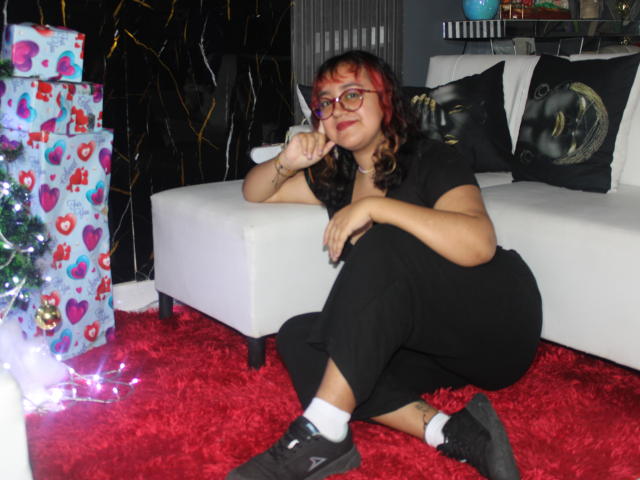 NamiNerona with red and black hair, glasses, black outfit, and black shoes, sitting on red carpet, holding finger to cheek, next to Christmas tree and gifts.