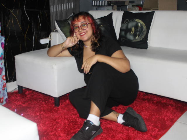 NamiNerona with red hair, glasses, and a smile, kneeling on a red rug, wearing black clothes, socks, and sneakers, leaning on a white couch with black and green pillows.