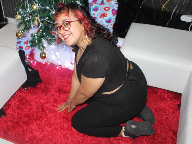 NamiNerona, curvy, black-tressed, red-haired, glasses-wearing, smiling, kneeling on red carpet, black outfit, butt up, Christmas tree with lights and ornaments in background, white furniture.